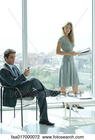 Office lobby, man sitting looking at cell phone while woman stands in background holding magazine View Large Photo Image Stock Image - Office lobby, man sitting looking at cell phone while woman stands in background holding magazine. Fotosearch