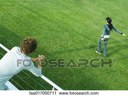 Stock Image - Teenage boy leaning on railing, watching teenage girl spinning on grass. Fotosearch
