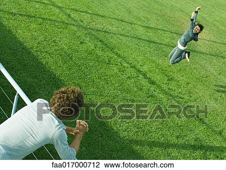 Stock Image - Teenage boy leaning on railing, watching teenage girl jumping on grass. Fotosearch