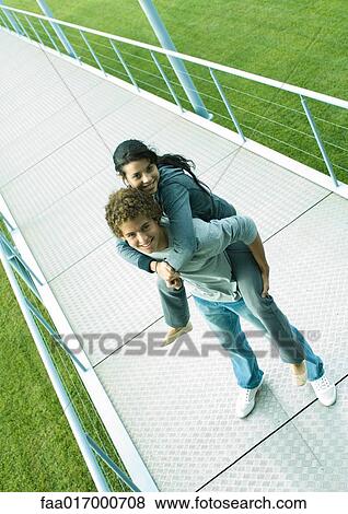 Stock Photo - Teenage couple, boy holding girl piggyback, standing on walkway smiling at camera. Fotosearch
