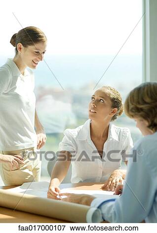 Stock Photo - Three adults gathered around blueprints in office. Fotosearch