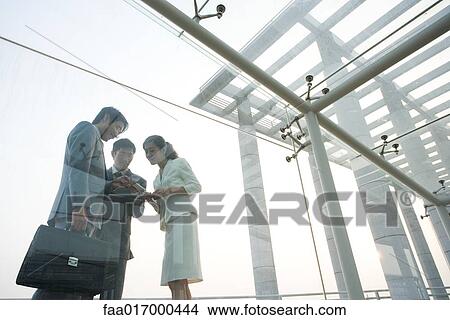 Picture - Three business executives standing in glass walkway, talking, low angle view. Fotosearch