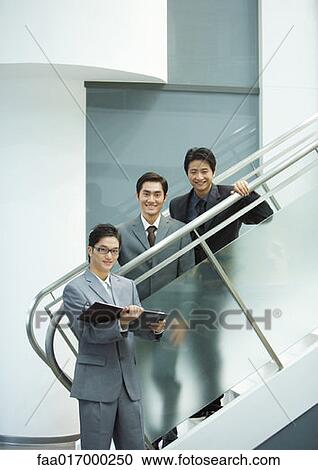 Stock Image - Three businessmen, two standing on stairs, smiling at camera. Fotosearch