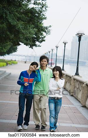 Picture - Three young adult friends walking by riverside, women making finger gestures. Fotosearch