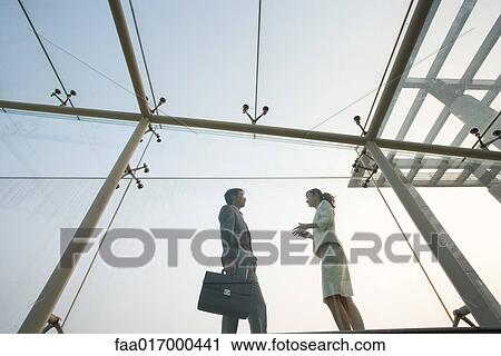 Stock Image - Two business executives standing in glass walkway, talking, low angle view. Fotosearch