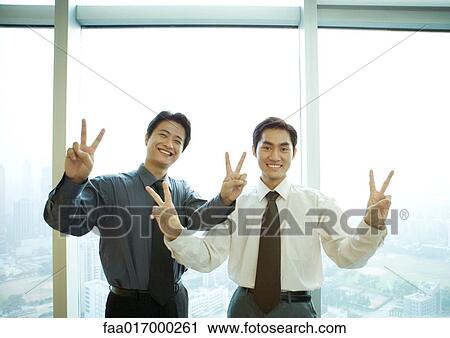 Two businessmen standing near window in high rise, making victory sign, smiling at camera View Large Photo Image Stock Image - Two businessmen standing near window in high rise, making victory sign, smiling at camera. Fotosearch