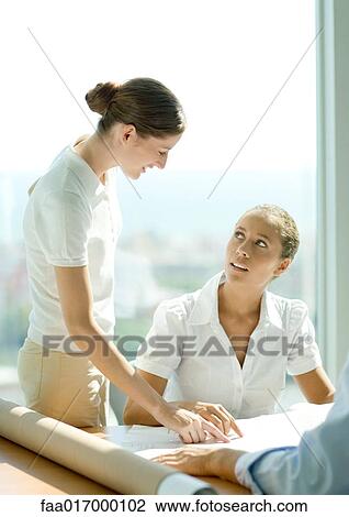 Stock Image - Two women discussing blueprints in office. Fotosearch