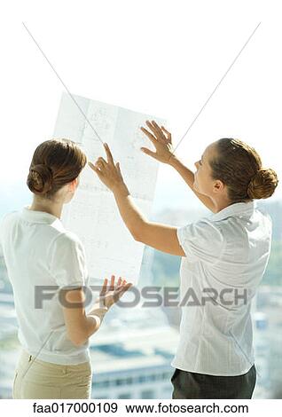 Stock Photo - Two women holding up blueprints against window, discussing, rear view. Fotosearch