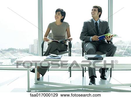 Stock Photo - Two young professional adults sitting in waiting room, smiling and looking out of frame. Fotosearch