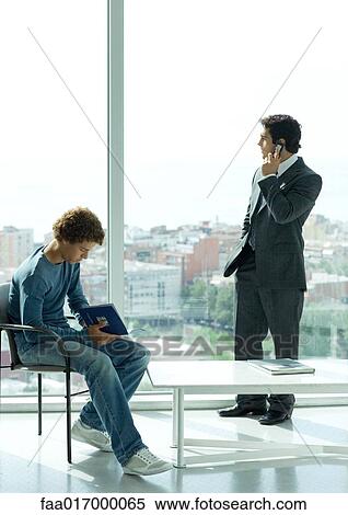 Stock Photography - Waiting room, casually dressed young man sitting in chair while businessman uses phone by window. Fotosearch
