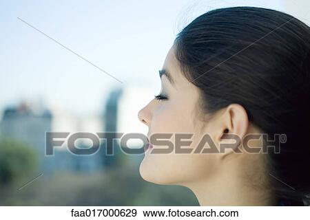 Stock Photo - Woman closing eyes, close-up, city in blurred background. Fotosearch