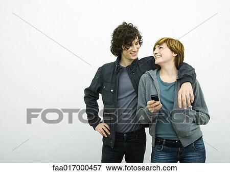 Young adult couple looking at each other, man with arm around woman, woman holding cell phone View Large Photo Image Stock Photo - Young adult couple looking at each other, man with arm around woman, woman holding cell phone. Fotosearch