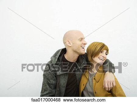 Stock Photo - Young adult couple looking out of frame, laughing, man with arm around woman. Fotosearch