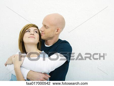 Stock Image - Young couple, man holding woman from behind, looking at each other. Fotosearch
