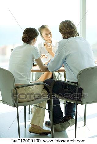 Stock Image - Young couple sitting across table from young female professional using phone. Fotosearch