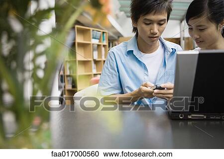 Stock Image - Young couple using laptop and cell phone together. Fotosearch