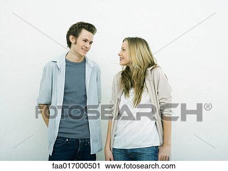 Stock Image - Young man and teenage girl standing side by side, smiling at each other. Fotosearch