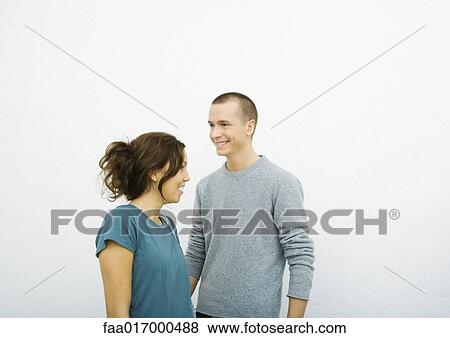 Stock Photo - Young man and young woman standing together, looking in opposite directions, smiling. Fotosearch