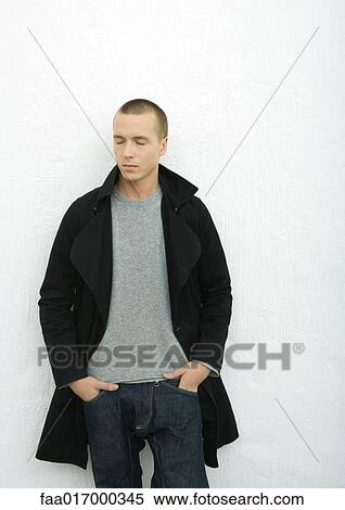 Stock Photography - Young man wearing coat, standing with hands in pockets and eyes closed, portrait. Fotosearch