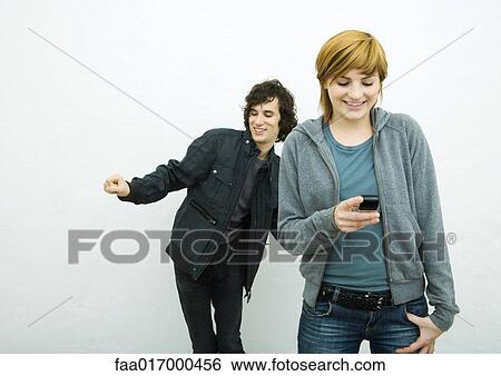 Stock Photograph - Young woman holding cell phone while young man behind her dances, white background. Fotosearch