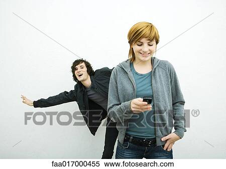 Stock Photography - Young woman holding cell phone while young man behind her dances, white background. Fotosearch