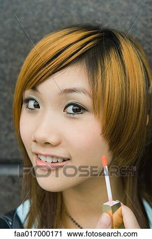 Stock Image - Young woman holding up lip gloss wand, smiling at camera. Fotosearch