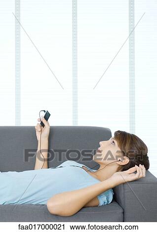 Stock Image - Young woman lying on couch, listening to MP3 player. Fotosearch