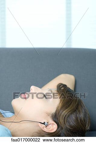 Stock Image - Young woman lying on couch, listening to earphones. Fotosearch