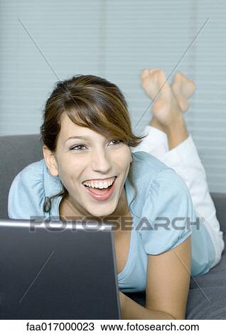 Stock Image - Young woman lying on couch, using laptop, laughing. Fotosearch