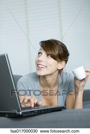 Young woman on couch, using laptop and holding coffee cup, looking up View Large Photo Image Stock Image - Young woman on couch, using laptop and holding coffee cup, looking up. Fotosearch