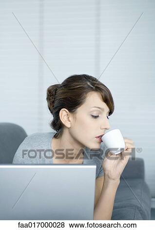 Stock Photo - Young woman on couch, using laptop and drinking coffee. Fotosearch