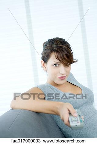 Stock Image - Young woman pointing remote control, smiling at camera. Fotosearch