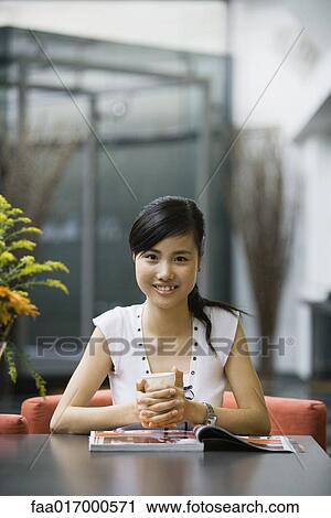 Stock Image - Young woman sitting with magazine and cup, smiling at camera. Fotosearch
