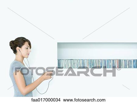 Picture - Young woman standing, listening to MP3 player, next to row of CDs, eyes closed. Fotosearch