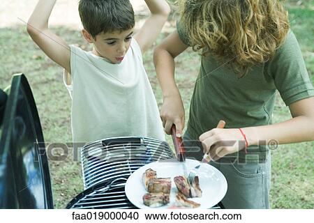 Stock Image - Boy cutting up meat on plate while second boy watches. Fotosearch