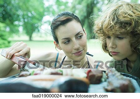 Boy looking at meats cooking on barbecue, woman glancing at him View Large Photo Image Picture - Boy looking at meats cooking on barbecue, woman glancing at him. Fotosearch