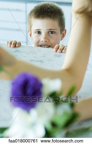 Stock Image - Boy looking over edge of counter, woman and flowers in blurred foreground. Fotosearch