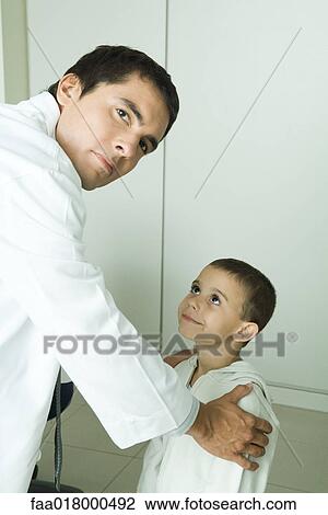 Stock Image - Boy looking up at doctor, doctor holding boy by shoulders, looking over shoulder at camera. Fotosearch