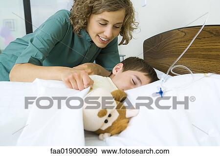 Stock Image - Boy lying in hospital bed asleep, nurse covering boy with blanket. Fotosearch