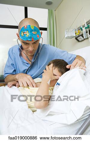 Boy lying in hospital bed holding head, doctor listening to boy with stethoscope View Large Photo Image Stock Photograph - Boy lying in hospital bed holding head, doctor listening to boy with stethoscope. Fotosearch