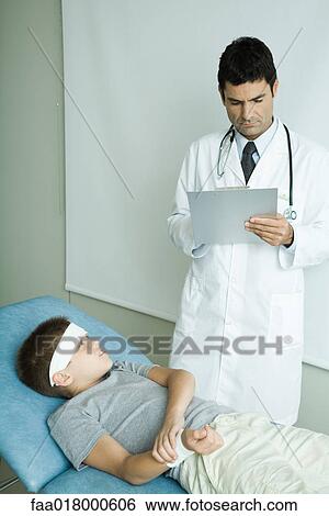 Boy lying on examination table with bandage on forehead, holding arm, doctor writing on clipboard View Large Photo Image Stock Photograph - Boy lying on examination table with bandage on forehead, holding arm, doctor writing on clipboard. Fotosearch