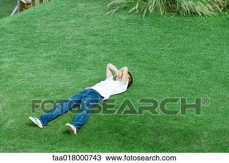Stock Image - Boy lying on grass, covering face with arms, full length. Fotosearch