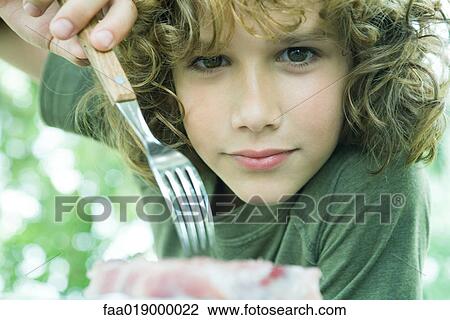 Stock Image - Boy picking up piece of meat with fork, looking at camera. Fotosearch