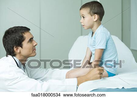 Boy sitting on examination table, doctor looking up at boy, profile View Large Photo Image Picture - Boy sitting on examination table, doctor looking up at boy, profile. Fotosearch