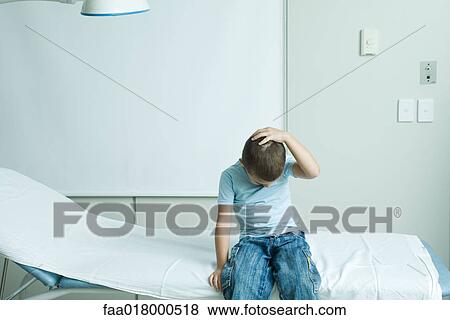 Stock Photo - Boy sitting on examination table, holding head. Fotosearch