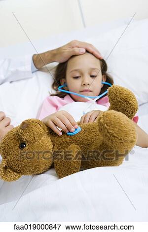 Stock Photo - Child holding toy stethoscope to teddy bear, doctor feeling child's forehead. Fotosearch