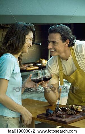 Stock Image - Couple standing in front of barbecue, clinking glasses of red wine and smiling at each other. Fotosearch