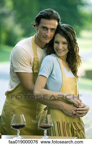 Stock Photograph - Couple wearing aprons, man standing behind woman with arms around her waist, portrait. Fotosearch