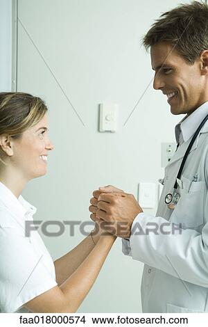 Picture - Doctor and woman standing face to face, holding hands and smiling at each other. Fotosearch