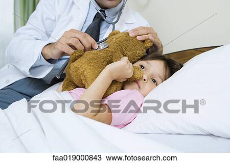 Stock Image - Doctor holding stethoscope to sick child's teddy bear. Fotosearch